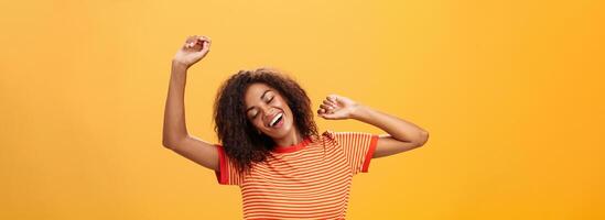 cintura para arriba Disparo de relajado y aliviado contento africano americano joven hembra estudiante en a rayas camiseta extensión brazos arriba clausura ojos desde deleite y sonriente teniendo genial siesta terminado naranja antecedentes foto