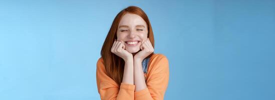 Silly cute happy redhead girlfriend smiling broadly close eyes dreamy squeez cheeks delighted asked date guy likes standing blue background rejoicing have fantastic lucky day triumphing photo