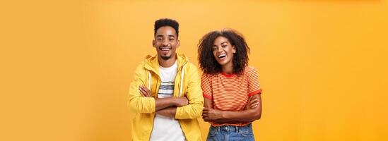 Two african american man and woman being best friends laughing out loud watching funny movie in cinema all dressed up in stylish outfit standing with hands crossed on chest and amused expression photo