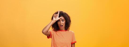 Girl thinks she loser. Portrait of gloomy bothered and displeased african american woman with afro hairstyle showing l word over forehead complaining feeling gloomy and unhappy over orange background photo