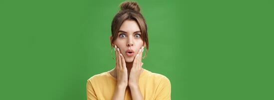 Portrait of impressed and shocked cute feminine woman in yellow t-shirt folding lips from excitement and interest touching cheeks surprised reacting to amazing rumor posing over green background photo