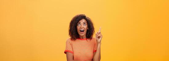 Waist-up shot of interested curious good-looking dark-skinned female in striped t-shirt talking asking question about curious star looking and pointing up with joy over orange background photo