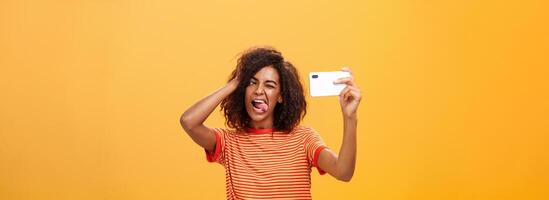 cintura para arriba Disparo de elegante confidente africano americano mujer con afro peinado en de moda camiseta tomando selfie en increíble nuevo Teléfono móvil pega fuera lengua jugando con pelo y parpadeo a teléfono inteligente foto