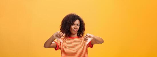 You loser and idea sucks. Dissatisfied and displeased young creative female artist with curly hairstyle frowning, sulking showing thumbd down in dislike gesture giving negative answer over orange wall photo