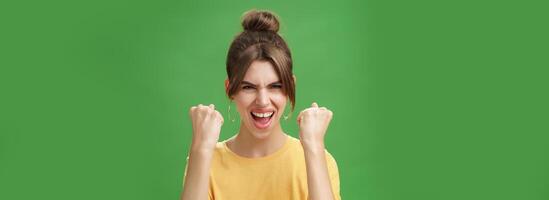 mujer con espíritu de ganador levantamiento apretado puños sonriente emocionado y apoyo aplausos siendo Listo para gimnasio ejercitado abucheos confidentes con grito mirando atrevido a cámara terminado verde pared foto