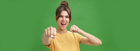 Cute female rebel in yellow t-shirt with gap teeth pulling fist towards camera as if showing fighting skills yelling daring and excited standing over green background smiling acting like boxer photo