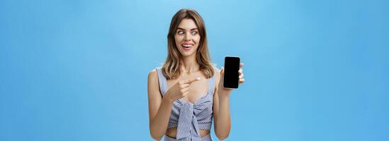 Portrait of charismatic astonished and happy woman with cute gapped teeth smiling axcited and surprised holding awesome smartphone pointing at device screen and looking at gadget amazed and delighted photo