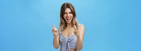 Charismatic joyful good-looking woman in trendy matching outfit winking joyfully making finger fun gesture at camera smiling broadly showing cute gapped teeth posing happy over blue wall photo