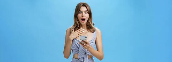 mujer sensación sorprendido y agradecido después leyendo inesperado Noticias desde teléfono inteligente prensado palma a cofre desde asombro goteante mandíbula y sonriente curioso a cámara, no puedo creer eso sucedió foto