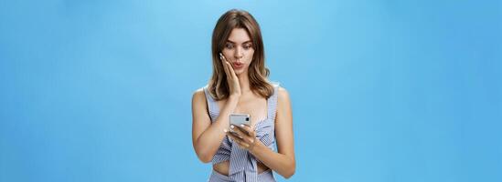 Woman learning shocking rumor receiving message in social network from friend reading news from smartphone folding lips staring excited at smartphone screen pressing palm to cheek from amazement photo