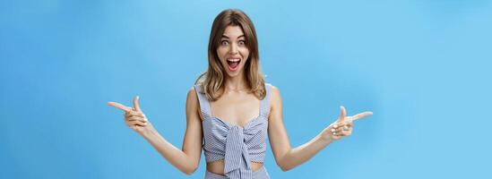 Amazed and surprised charismatic attractive woman raising hands pointing sideways left and right with impressed overwhelmed expression, smiling broadly liking both sides of copy space over blue wall photo