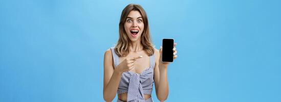 emocionado contento mujer con brechado dientes finalmente compró marca nuevo teléfono inteligente participación dispositivo en mano señalando a Teléfono móvil pantalla demostración frio aplicación sonriente en general desde alegría en contra azul pared foto