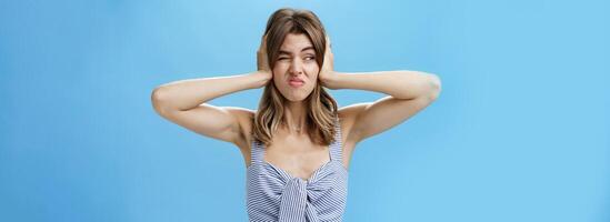 Girl feeling irritated by loud awful noise pressing hands to ears not hear annoying sound of drill smirking and frowning, squinting looking left displeased and disturbed, posing over blue background photo