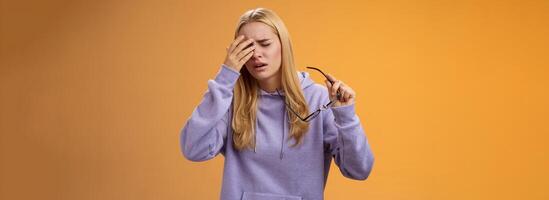 Tired dizzy cute blond girl taking off glasses touching forehead feel sick unwell suffering headache painful migraine standing bothered uncomfortable orange background in hoodie photo