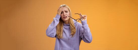 Upset gloomy bothered blond girl take-off glasses cringing grimacing feel unwell suffering headache standing dizzy touching forehead painful migraine bothering working woman, orange background photo