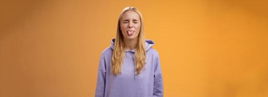 Playful amused charismatic young funny blond girl having fun close eyes showing tongue rebellious fool around unbothered enjoying carefree sunny spring days standing orange background photo