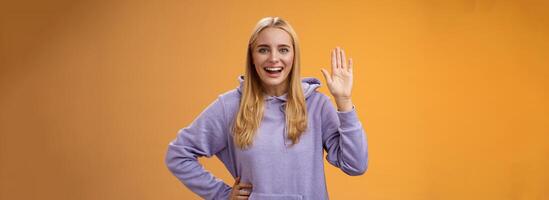 Friendly energized helpful charming female student say hello newcomers waving raised palm hey hi gesture greeting welcoming happily, standing orange background smiling broadly photo