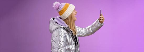 Amused carefree attractive caucasian blond girl in silver winter jacket hat extend arm holding smartphone recording own yell open mouth wide close eyes fool around mimicking funny faces selfie photo
