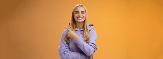 Charismatic modern millennial cute blond woman in hoodie smiling happily discussing cool new promo pointing right recommend check out visit interesting store near corner, orange background photo
