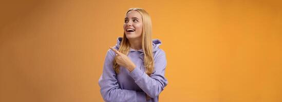 Amused carefree joyful blond european woman in hoodie pointing looking right pleased smiling happily enjoying interesting fascinating peromance making choice in store shopping, orange background photo