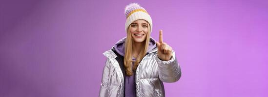 Excited cheerful fair-haired european girl in winter hat silver shiny jacket show number one index fingers give suggestion advice smiling broadly talking happily, purple background photo