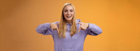 Amused joyful nice blond girlfriend pointing down present cool new product smiling broadly recommending try check out standing orange background happily grinning in hoodie photo