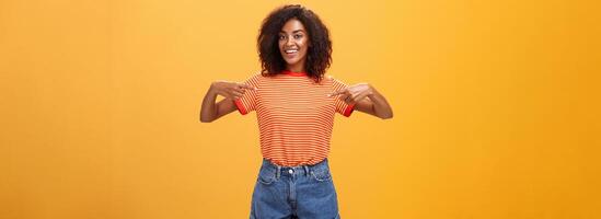 Oye recoger yo yo tu niña mirando para. retrato de encantador de apariencia amistosa ambicioso de piel oscura hembra con afro peinado señalando a cofre con orgullo y alegre posando en contra naranja antecedentes foto