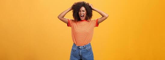 yo odio vida siendo cruel. Deprimido africano mujer en rabia y furia tracción pelo fuera de cabeza Gritando y gritando con cerrado ojos perdiendo templar teniendo malo día odiando todo terminado naranja pared foto