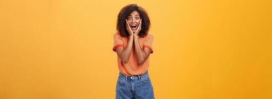 Impressed talkative african american woman finding out hot details of rumor being excited, thrilled leaning face on palms, smiling broadly with amused expression listening carefully over orange wall photo