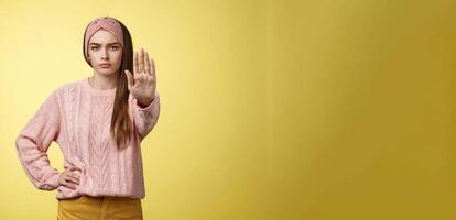 Stop it. Serious-looking confident focused young cute girl extending arm hold, tresspass gesture, frowning displeased, standing bossy denying, forbidding pass, unwilling let-through over yellow wall photo