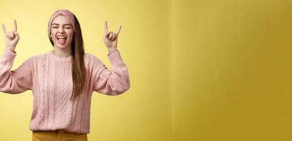 Cute heavy metal lover showing rock roll symbol sticking tongue amused and happy fooling around listening favourite music posing excited and pleased against yellow background in knitted outfit photo