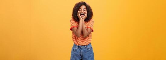 Woman happy and delighted with great result after curing acne with skincare products touching cheeks smiling happily with closed eyes feeling pleased and amused over orange background photo