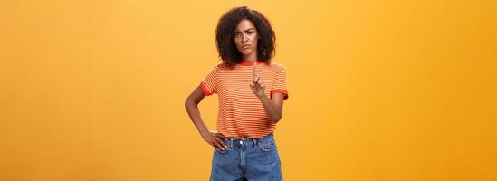 You better not. Dissatisfied bossy serious-looking african american curly-haired female head manager in t-shirt frowning shaking index finger in forbid or stop gesture scolding dog over orange wall photo