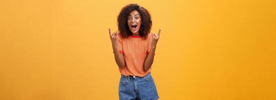 Stylish african american woman being delighted and excited before cool concert showing rock n roll gesture with both hands screaming from satisfaction and happiness cheering over orange background photo