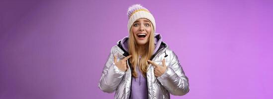 Surprised happy smiling joyful attractive woman pointing herself satisfied picked was chosen winner standing excited in silver shiny jacket winter hat cannot believe own luck, purple background photo