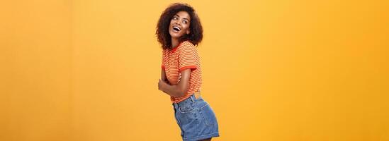 Hey wanna hang out. Sensual and flirty carefree stylish dark-skinned woman with curly hair standing in profile turning left and posing with happy cute expression smiling and seducing over orange wall photo