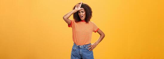 Arrogant and confident woman not talk with losers. Mocking self-assured stylish african american female with curly hair in trendy clothes frowning scolding nerd, showing letter l on forehead photo