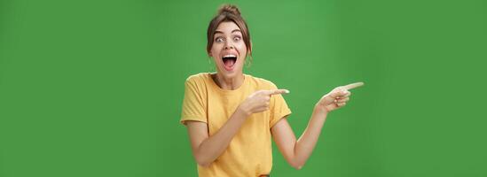 Waist-up shot of enthusiastic and charismatic impressed happy girl laughing and smiling broadly pointing left at funny hilarious thing reacting surprised and entertained, posing over green background photo