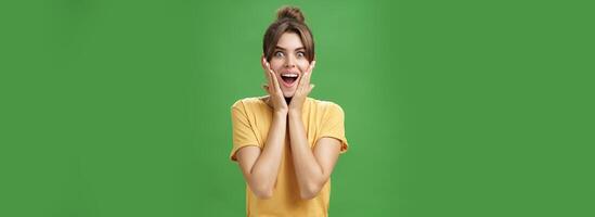 Indoor shot of excited and surprised energized attractive glamorous female in yellow t-shirt touching cheeks from amazement smiling broadly astonished posing over green background photo