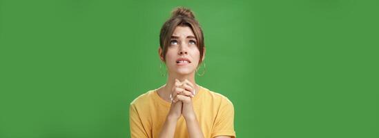 Intense nervous and faithful good-looking young woman clenching hands together over chest looking up and biting lower lip while praying god help, standing hopeful over green background photo