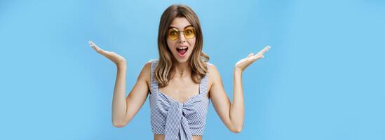 cintura para arriba Disparo de carismático linda joven mujer en Gafas de sol levantamiento manos oblicuo en despistado y inseguro gesto encogiéndose de hombros con abrió boca y tonto sonrisa siendo sin molestarse, posando en contra azul pared foto