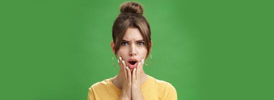 Shocked woman reacting to pimple looking disturbed and displeased in mirror holding hands pressed to cheeks open mouth and frowning being disappointed and upset over green background photo