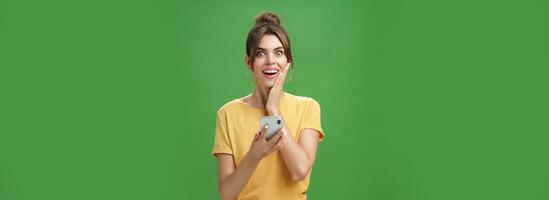 Surprised touched and impressed beautiful european girl with diastema smiling broadly touching cheek from amazement and joy holding smartphone looking happy at camera after reading cool news photo