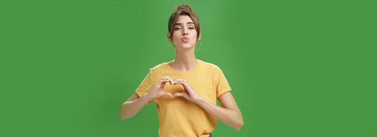 Sensual charming woman in yellow t-shirt and combed hairstyle in yellow t-shirt showing heart gesture against chest folding lips to give kiss or mwah tender and loving over green background photo