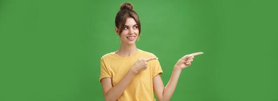 Waist-up shot of good-looking friendly and feminine female coworker in yellow t-shirt showing newbie office pointing and looking left with cute happy smile posing against green background photo