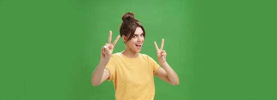 Indoor shot of enthusiastic excited and happy daring girl with combed hair tattoo and cute diasdema showing peace signs bending backwards standing in cool energized pose over green background photo