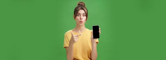 Portrait of intrigued woman cannot wait use new smartphone holding phone and pointing at cellphone screen folding lips with excitement and interest presenting new device model in store over green wall photo