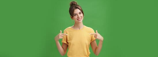 Calm and polite friendly-looking skillful young female coworker in yellow t-shirt pointing at herself self-assured tilting head and smiling at camera wanting participate in event over green background photo