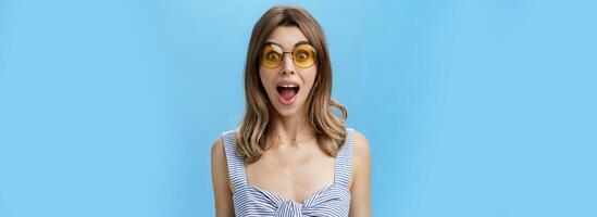 Waist-up shot of excited surprised and emotive charismatic caucasian woman in trendy sunglasses opening mouth from amazement and joy gazing at camera impressed and astonished over blue background photo