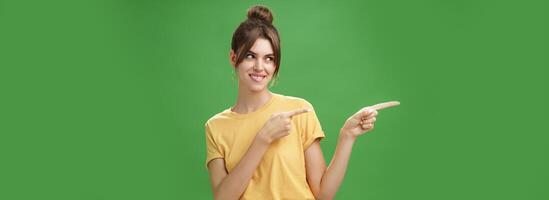 Waist-up shot of charismatic happy and carefree charming woman in yellow t-shirt pointing and looking left enthusiastic and pleased smiling cheerfully posing against green background delighted photo